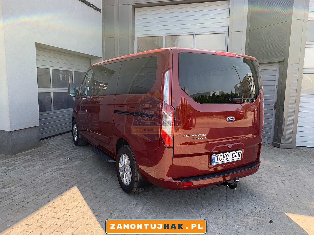 Zdjęcie przedstawia Forda Tourneo Custom z zamontowanym hakiem holowniczym wypinanym pionowo brink, zdjęcie zostało zrobione od tyłu z lewej strony na tle serwisu zamontujhak.pl